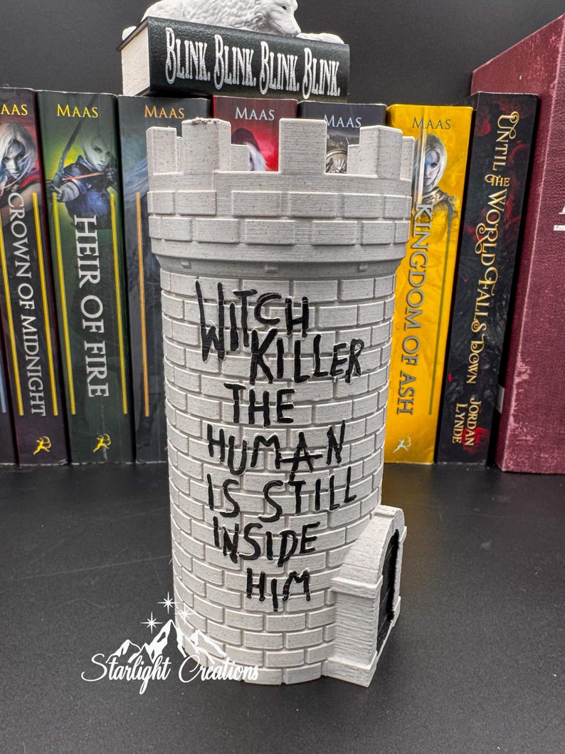 Puede incluir: Un objeto decorativo gris en forma de castillo con el texto "Witch Killer The Human Is Still Inside Him". El objeto tiene una parte superior almenada y una peque&ntilde;a puerta arqueada. Hay libros en el fondo.