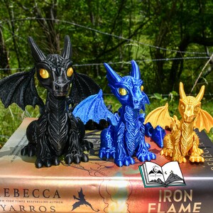 May include: Three dragon figurines in black, blue, and gold, each with detailed wings and large eyes. The dragons are sitting on a book with the title "Iron Flame". The background is a blurred view of green foliage.
