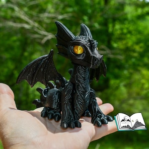 May include: A black dragon figurine with yellow eyes and detailed wings, held in a hand. The dragon has a textured surface and is posed with its wings spread. A small book with a mountain and moon logo is in the corner.