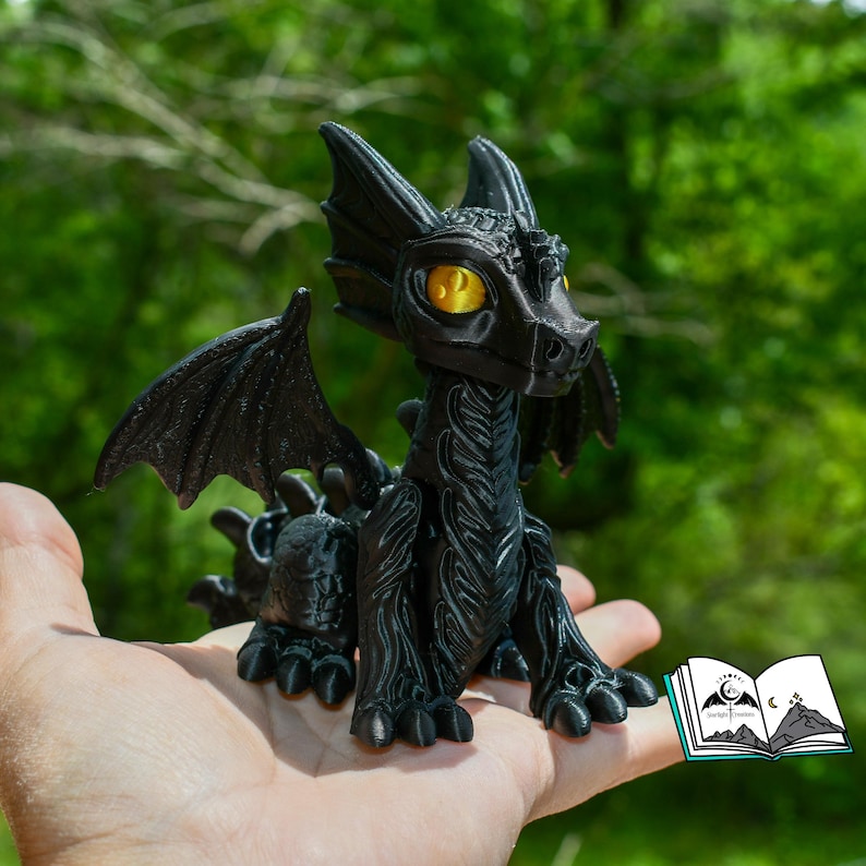 May include: A black dragon figurine with yellow eyes and detailed wings, held in a hand. The dragon has a textured surface and is posed with its wings spread. A small book with a mountain and moon logo is in the corner.