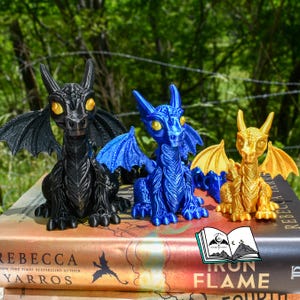 May include: Three dragon figurines in black, blue, and gold, with detailed wings and yellow eyes, sit atop books. The dragons are posed in a sitting position. The background is a blurred view of green trees.