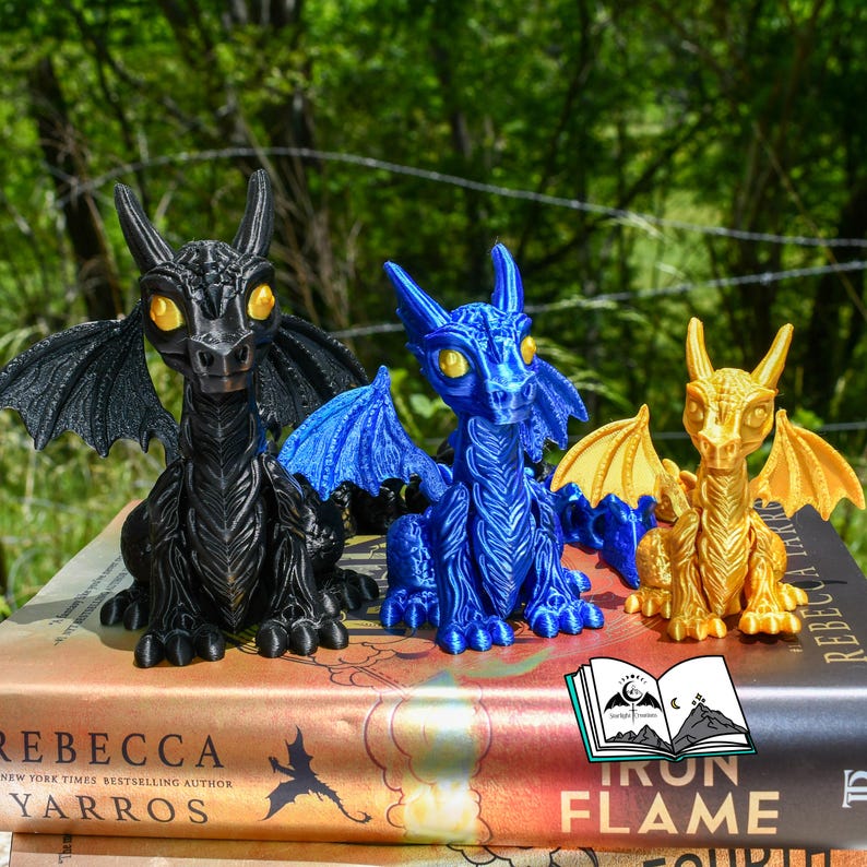 May include: Three dragon figurines in black, blue, and gold, with detailed wings and yellow eyes, sit atop books. The dragons are posed in a sitting position. The background is a blurred view of green trees.
