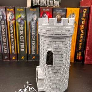 Puede incluir: Una torre de castillo gris impresa en 3D con una ventana rectangular y detalles de ladrillo. La torre est&aacute; frente a una fila de libros con la palabra "MAAS" en los lomos. Una peque&ntilde;a figura de lobo blanco descansa sobre un libro que dice "BLUNK BLUNK BLINK BLINK". El texto "Starlight Creations" est&aacute; en la parte inferior.