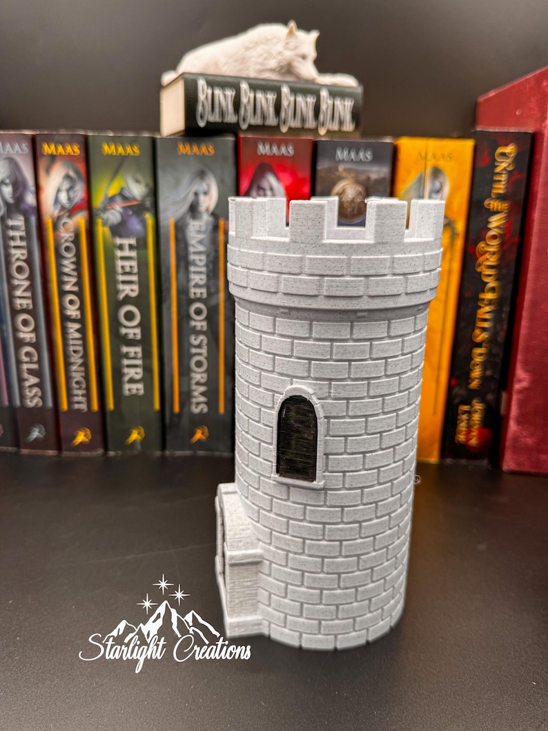 Puede incluir: Una torre de castillo gris impresa en 3D con una ventana rectangular y detalles de ladrillo. La torre est&aacute; frente a una fila de libros con la palabra "MAAS" en los lomos. Una peque&ntilde;a figura de lobo blanco descansa sobre un libro que dice "BLUNK BLUNK BLINK BLINK". El texto "Starlight Creations" est&aacute; en la parte inferior.