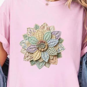 Può includere: Una t-shirt a maniche corte rosa chiaro con una grafica centrale raffigurante un fiore a più strati con petali in vari colori pastello e motivi, tra cui quadretti vichy, fiori e texture a tinta unita, indossata sotto una giacca di jeans blu.