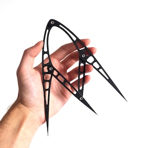 May include: A black metal divider tool with pointed ends and a curved design. The tool has multiple holes and is held in a person's hand against a white background. The tool is used for measuring and drawing circles.