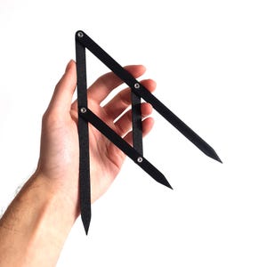 May include: A black proportional divider tool, held against a white background. The tool has three pivot points and pointed ends. The tool is used for scaling measurements in art and design.
