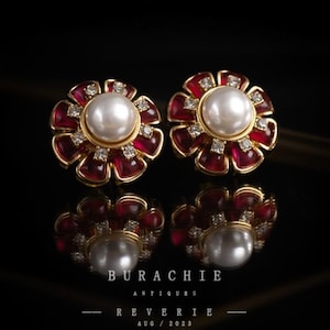 May include: A pair of flower-shaped stud earrings. Each earring features a large, white pearl center, surrounded by red petals and small, sparkling accents, set in a gold-toned metal. The earrings are displayed on a reflective surface.