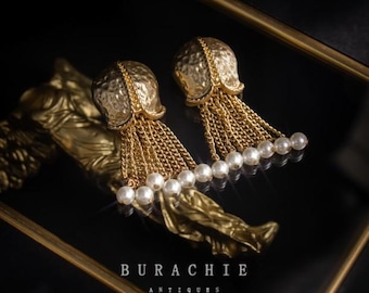 May include: A pair of gold-colored earrings with a textured, tulip-shaped top and dangling chains. Each chain ends with a row of white pearls. The earrings are displayed on a gold-colored decorative object. The text "BURACHIE ANTIQUES REVERIE AUG/2023" is visible.