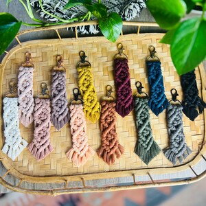 Puede incluir: Una colección de 12 llaveros de macramé de colores con flecos. Los llaveros se muestran en una bandeja tejida.