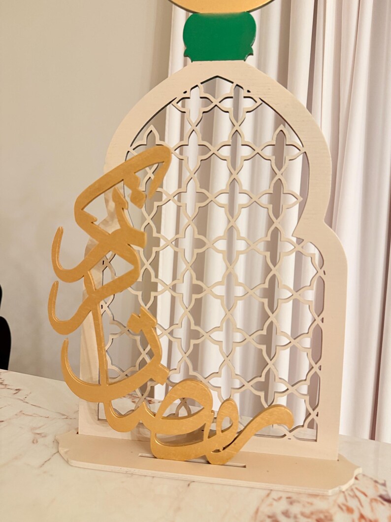 Puede incluir: Una pieza decorativa de madera con un dise&ntilde;o de ventana arqueada con un patr&oacute;n geom&eacute;trico. Caligraf&iacute;a &aacute;rabe dorada est&aacute; superpuesta frente a la ventana. La pieza est&aacute; pintada en tonos neutros con un acento verde.