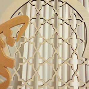 Puede incluir: Panel decorativo de madera con un dise&ntilde;o de celos&iacute;a geom&eacute;trica. El panel es de color beige claro con un acento dorado y una parte superior verde. El intrincado patr&oacute;n recortado es visible contra un fondo de cortina blanca.