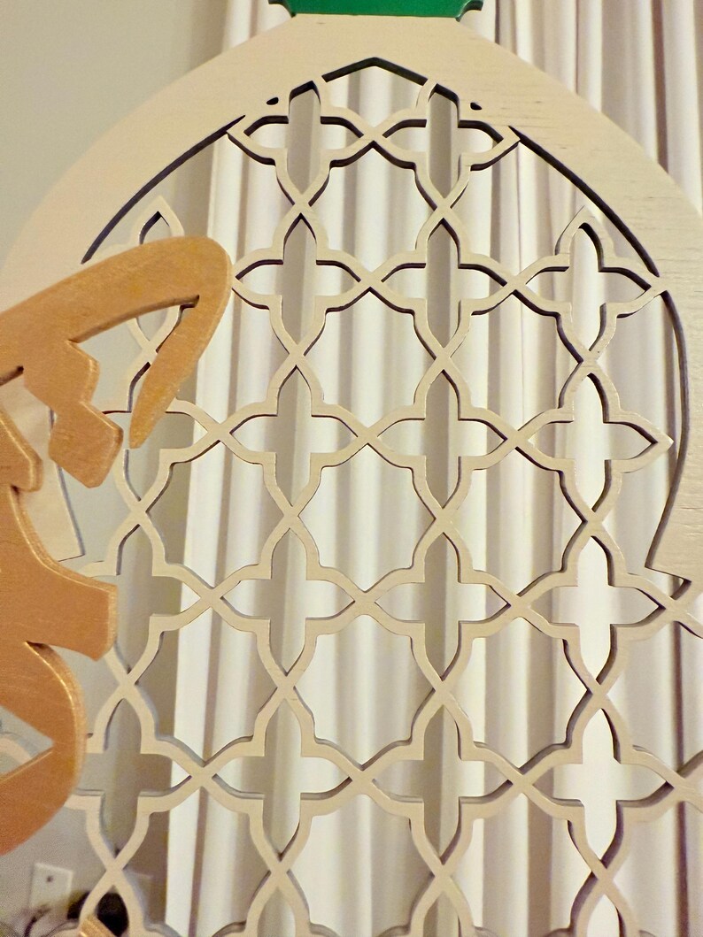 Puede incluir: Panel decorativo de madera con un dise&ntilde;o de celos&iacute;a geom&eacute;trica. El panel es de color beige claro con un acento dorado y una parte superior verde. El intrincado patr&oacute;n recortado es visible contra un fondo de cortina blanca.
