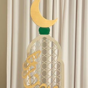 Puede incluir: Decoraci&oacute;n de madera para el Ramad&aacute;n. Presenta una luna creciente dorada sobre una base verde, encima de un arco crema con un dise&ntilde;o de celos&iacute;a. Caligraf&iacute;a &aacute;rabe dorada en la parte delantera. Fondo de cortina blanca.