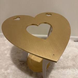 May include: A gold heart-shaped table with a white base and a smaller shelf. The table has three cut-out hearts on the top.