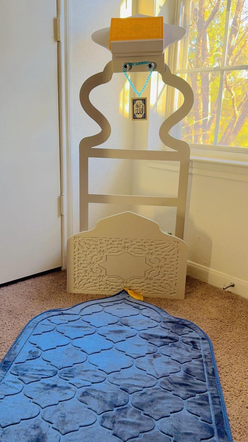 May include: A white wooden prayer stand with a blue prayer rug. The stand has a decorative design and a small shelf on top. The rug has a geometric pattern. The stand has a small tag with the word "Allah" written on it.