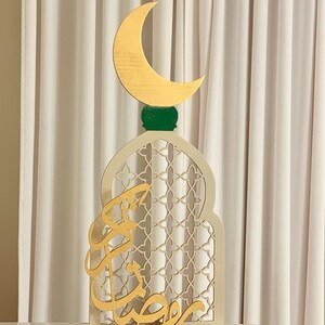 Puede incluir: Decoraci&oacute;n de madera para el Ramad&aacute;n. Presenta una luna creciente dorada sobre una base verde, sobre un arco crema con un dise&ntilde;o de celos&iacute;a. Caligraf&iacute;a &aacute;rabe dorada en la parte delantera. La decoraci&oacute;n est&aacute; sobre una superficie de m&aacute;rmol.