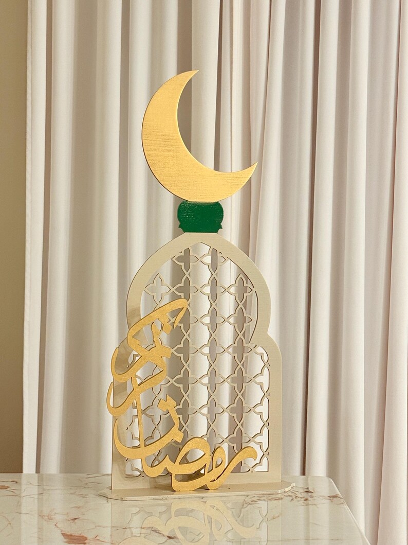 Puede incluir: Decoraci&oacute;n de madera para el Ramad&aacute;n. Presenta una luna creciente dorada sobre una base verde, sobre un arco crema con un dise&ntilde;o de celos&iacute;a. Caligraf&iacute;a &aacute;rabe dorada en la parte delantera. La decoraci&oacute;n est&aacute; sobre una superficie de m&aacute;rmol.