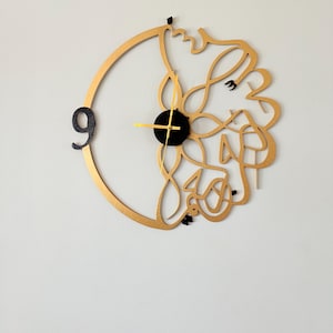 May include: A gold-colored wall clock with Arabic calligraphy design. The clock features a black center with gold hands and the number 9 in black. The design is abstract and decorative, suitable for home decor.