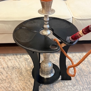 May include: A black hookah table with a silver metal hookah. The table has a round top with a cutout for the hookah and a curved leg design. The hookah has a brown bowl and a red and gold hose.