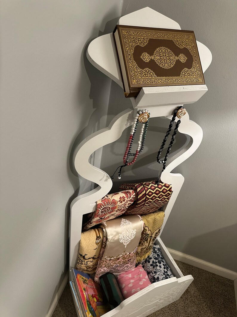 May include: White wooden prayer shelf with a brown book, two prayer beads, and a stack of prayer rugs. The shelf has a drawer with a book and other items inside.