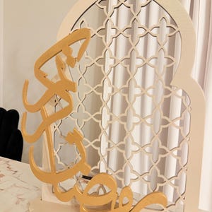 Puede incluir: Un letrero decorativo de madera para el Ramad&aacute;n. El letrero presenta un arco color crema con un dise&ntilde;o de celos&iacute;a geom&eacute;trica. Caligraf&iacute;a &aacute;rabe dorada est&aacute; superpuesta al arco. La parte superior del arco es verde. El letrero est&aacute; dise&ntilde;ado para colocarse sobre una superficie plana.