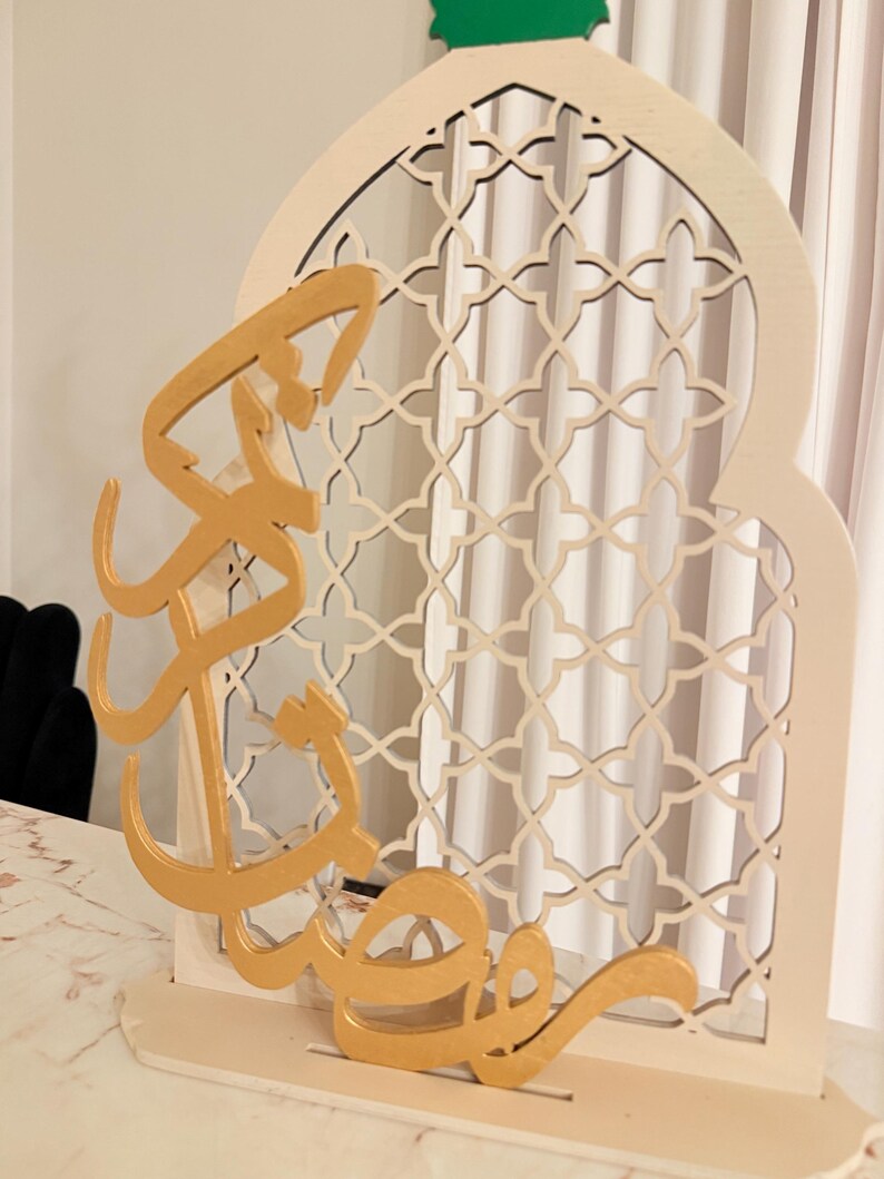 Puede incluir: Un letrero decorativo de madera para el Ramad&aacute;n. El letrero presenta un arco color crema con un dise&ntilde;o de celos&iacute;a geom&eacute;trica. Caligraf&iacute;a &aacute;rabe dorada est&aacute; superpuesta al arco. La parte superior del arco es verde. El letrero est&aacute; dise&ntilde;ado para colocarse sobre una superficie plana.