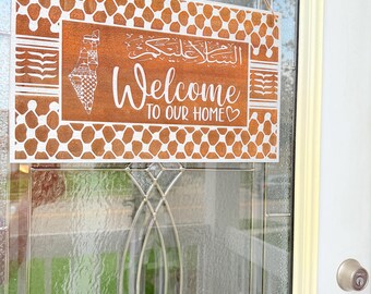 May include: A wooden welcome sign with a brown background and white geometric patterns. The sign says "Welcome to our home" with Arabic calligraphy above the text and a heart symbol after the text.