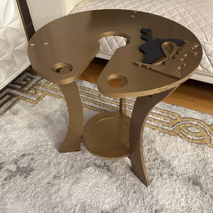 May include: A gold-colored metal table with a unique design. The table has a round top with a cutout in the shape of a U. The table has three legs that are curved and extend from the bottom of the table top.