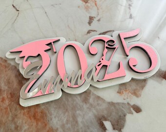 May include: Pink and white wooden sign with the year 2025 and the word "Emma" in silver lettering. The number 2 is decorated with a graduation cap.
