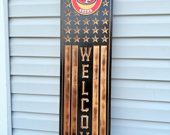 Puede incluir: Un letrero de bienvenida de madera con el logotipo de los 49ers de San Francisco. El letrero tiene un fondo negro con estrellas doradas y rayas verticales. La palabra "WELCOME" está tallada en letras grandes.