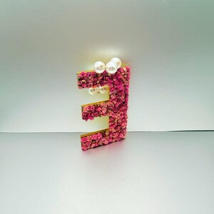 May include: A decorative letter "E" with a gold frame, covered in pink and peach-colored flowers. The letter is adorned with white pearl accents, creating a floral and elegant design. The background is a light gray.