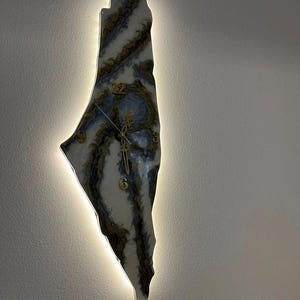 May include: A white and blue resin clock shaped like the outline of Israel with gold accents. The clock is illuminated with a white light.