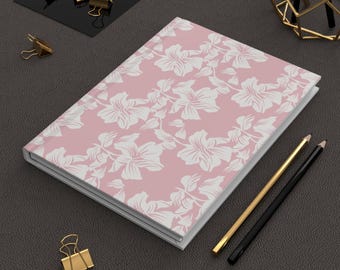 Può includere: Un diario con copertina rigida con un motivo floreale rosa. Il design floreale bianco è posto su uno sfondo rosa. Sono visibili anche matite dorate e nere e fermagli dorati.