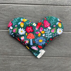 May include: A green heart-shaped pillow with a colorful floral and mushroom pattern. The pillow has a white tag with the letters "K.P." on it.