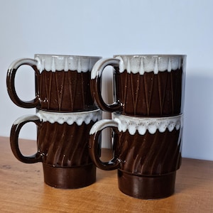 May include: Four stacked brown ceramic mugs with white trim. Each mug has a large handle and a textured, geometric design. The mugs are arranged on a wooden surface against a white background.