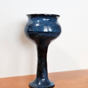 Puede incluir: Un jarrón de cerámica hecho a mano con un esmalte azul oscuro. El jarrón tiene una parte superior bulbosa, un tallo delgado y una base acampanada. El color azul oscuro tiene una textura moteada, lo que añade interés visual a la pieza. El jarrón se exhibe sobre una superficie de madera.