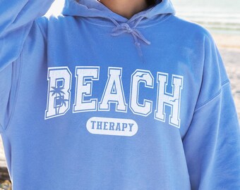 May include: A light blue hoodie with the text "Beach Therapy" in white lettering. The hoodie has a large pocket on the front and a drawstring hood.