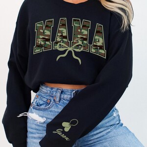 K&ouml;nnte beinhalten: Schwarzer, kurzer Pullover mit einem Tarnmuster-Druck "MAMA" und einer gr&uuml;nen Schleife. Der Pullover hat ein personalisiertes Detail am &Auml;rmel mit dem Namen "Jason" und einem gr&uuml;nen Herzen.