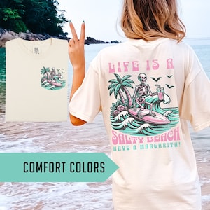K&ouml;nnte beinhalten: Ein wei&szlig;es T-Shirt mit einem Aufdruck eines Skeletts, das surft und eine Margarita h&auml;lt. Der Text auf dem Shirt lautet "Life is a Salty Beach Have a Margarita!" Das Shirt wird an einem Model gezeigt, das am Strand steht.