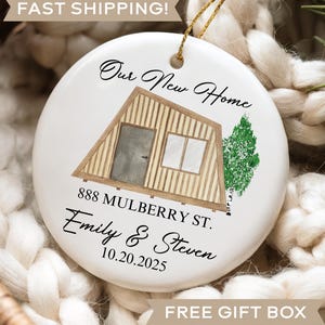 May include: A white ceramic ornament with a gold string for hanging. The ornament features a drawing of a house and the text "Our New Home," address, names, and a date. The ornament is nestled in a woven basket.