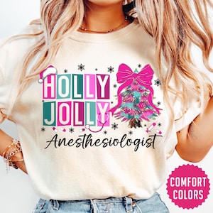May include: A white t-shirt with a Christmas tree design and the text "Holly Jolly Anesthesiologist". The tree is decorated with pink and green ornaments and a pink bow. The text is in pink, green, and blue.