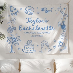 May include: A white fabric tapestry with blue illustrations of a palm tree, cake, champagne flutes, and other celebratory items. The text "Taylor's Bachelorette San Diego, California July 2025" is printed on the tapestry.