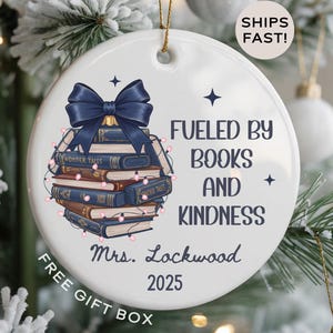 May include: A white ceramic Christmas ornament with a navy blue bow and a stack of books. The ornament reads "Fueled by Books and Kindness" and "Mrs. Lockwood 2025". The ornament is decorated with string lights.