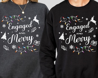 May include: Two sweatshirts, one dark heather and one black, each with the text "Engaged and Merry" in white script. The design includes a string of colorful lights, snowflakes, a Christmas tree, a gift, and a reindeer.