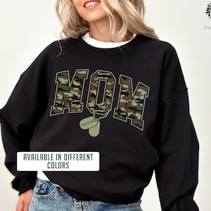 May include: Black sweatshirt with a camouflage print of the word "MOM" and a dog tag with the name "TREVOR" printed on it. The text "AVAILABLE IN DIFFERENT COLORS" is printed below the design. The sweatshirt is being modeled by a person wearing blue jeans.