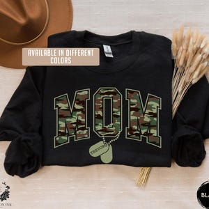 May include: Black sweatshirt with a camouflage print of the word "MOM" and a dog tag with the name "TREVOR" printed on it. The text "AVAILABLE IN DIFFERENT COLORS" is printed on the top left corner of the sweatshirt.