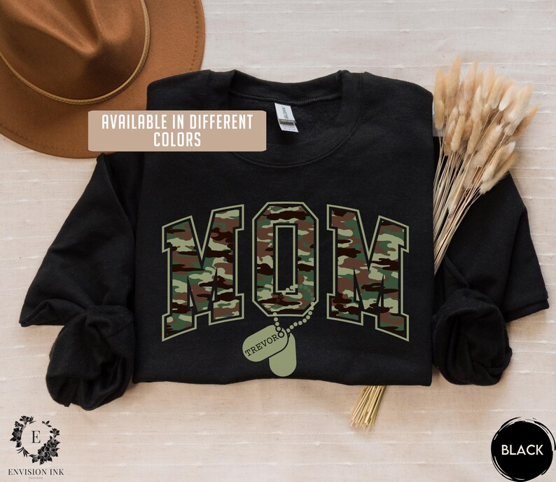 May include: Black sweatshirt with a camouflage print of the word "MOM" and a dog tag with the name "TREVOR" printed on it. The text "AVAILABLE IN DIFFERENT COLORS" is printed on the top left corner of the sweatshirt.
