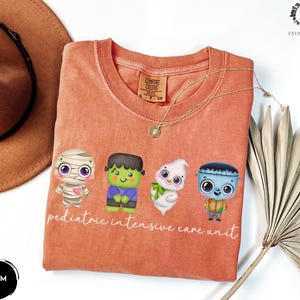 Pode incluir: Uma camiseta laranja claro com um gr&aacute;fico de quatro monstros de desenho animado fofinhos. O texto "pediatric intensive care unit" &eacute; impresso abaixo dos monstros.