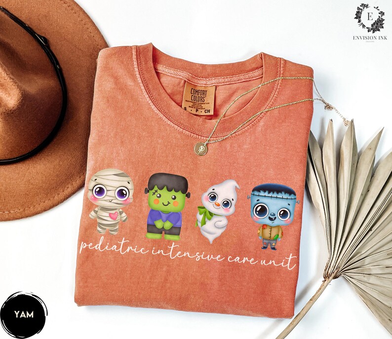 Pode incluir: Uma camiseta laranja claro com um gr&aacute;fico de quatro monstros de desenho animado fofinhos. O texto "pediatric intensive care unit" &eacute; impresso abaixo dos monstros.